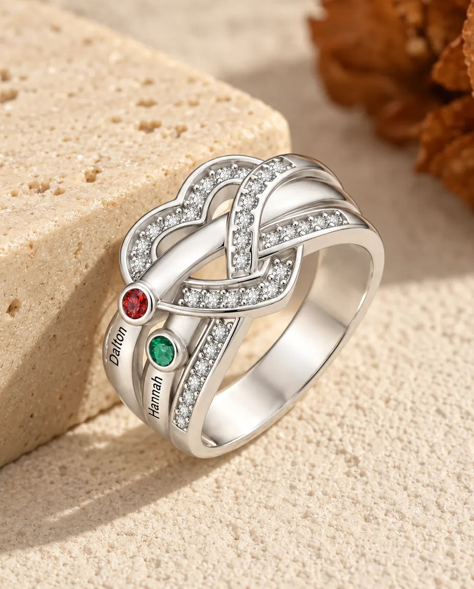 personalized heart knot birthstone ring with 2 names and 2 round birthstones, rhodium plated, on travertine