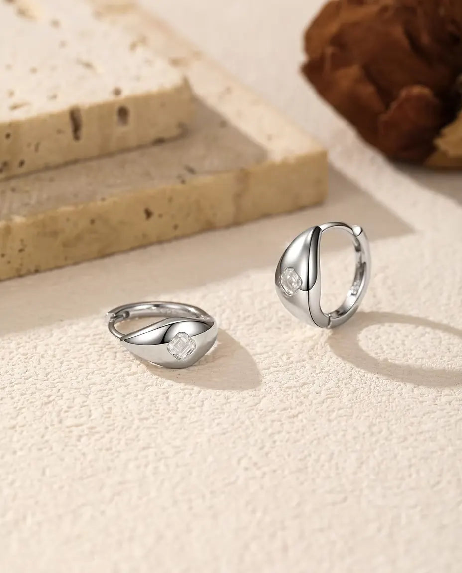 Asscher Cut Moissanite Huggie Hoop Earrings in 925 Sterling Silver - pair on a cream textured surface, one lying flat and one standing