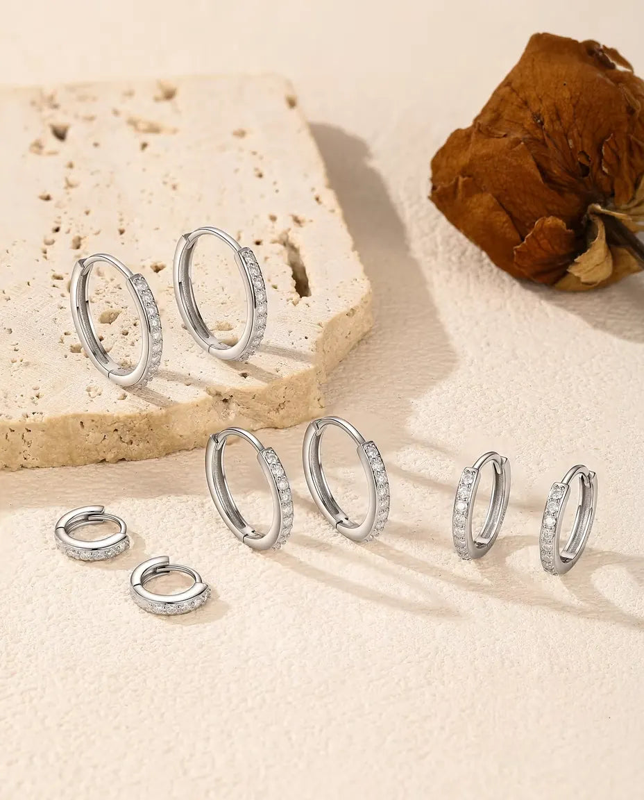 Moissanite Huggie Hoop Earrings in 925 Sterling Silver - all five sizes arranged on a cream surface by a travertine slab