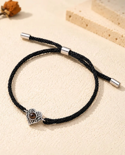 Personalized photo projection bracelet with a silver tone angel wing heart on a black rope cord, lying on cream plaster