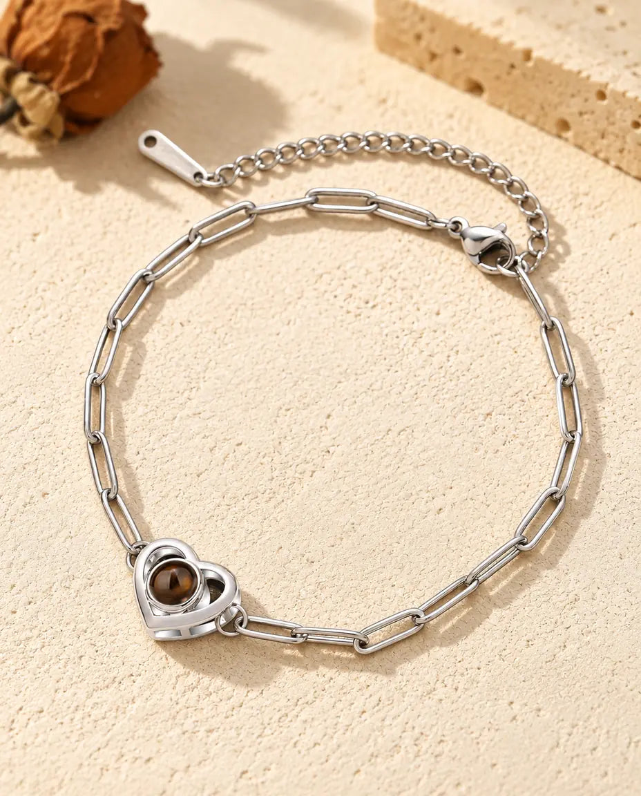 Personalized photo projection bracelet in silver tone with a double heart charm on a paperclip chain, lying on cream plaster