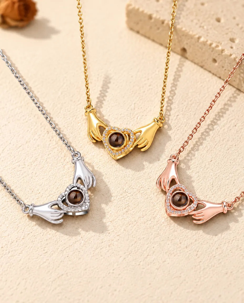 Three colors of the hands holding heart photo projection necklace in a row on cream plaster