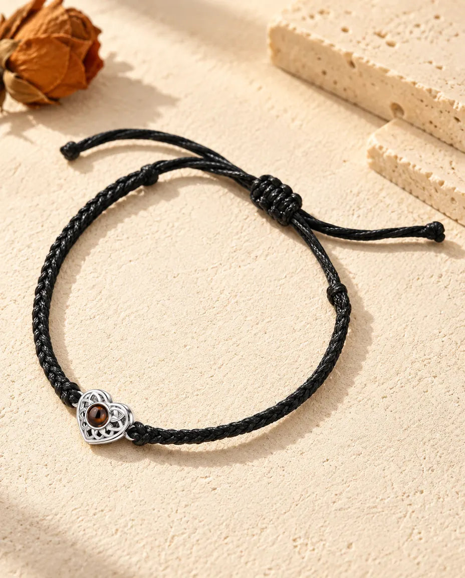 Personalized photo projection bracelet with a silver tone openwork heart charm on a black thin braided cord, lying on cream plaster
