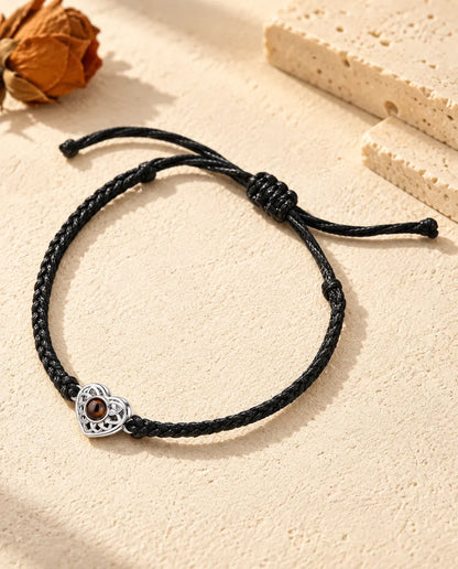Personalized photo projection bracelet with a silver tone openwork heart charm on a black thin braided cord, lying on cream plaster