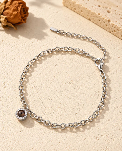 Personalized photo projection bracelet in silver tone with a tiny pave circle charm on a fine chain, lying on cream plaster