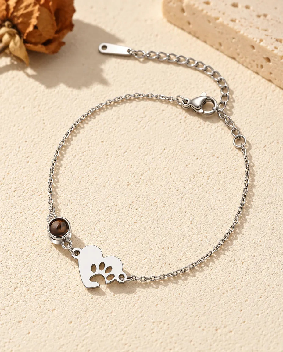 Personalized photo projection bracelet with a silver tone paw print heart tag and projection stone on a fine chain, lying on cream plaster