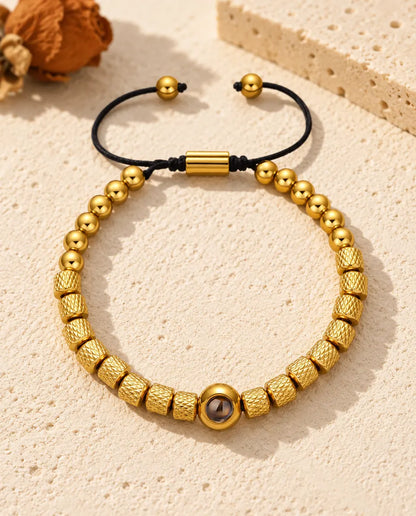 Personalized photo projection bead bracelet in gold tone with textured barrel beads and a sliding cord, on cream plaster
