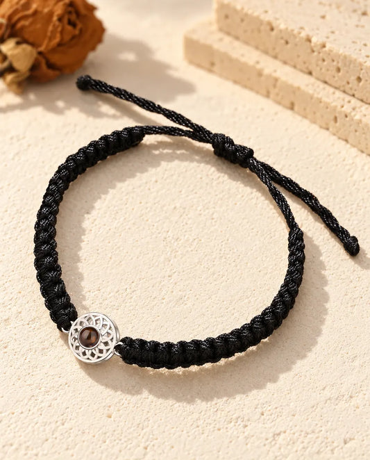 Personalized photo projection bracelet with a silver tone openwork round charm on a black braided cord, lying on cream plaster