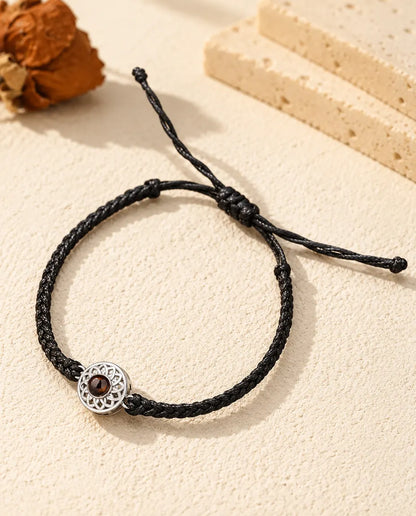 Personalized photo projection bracelet with a silver tone openwork round charm on a black thin braided cord, lying on cream plaster