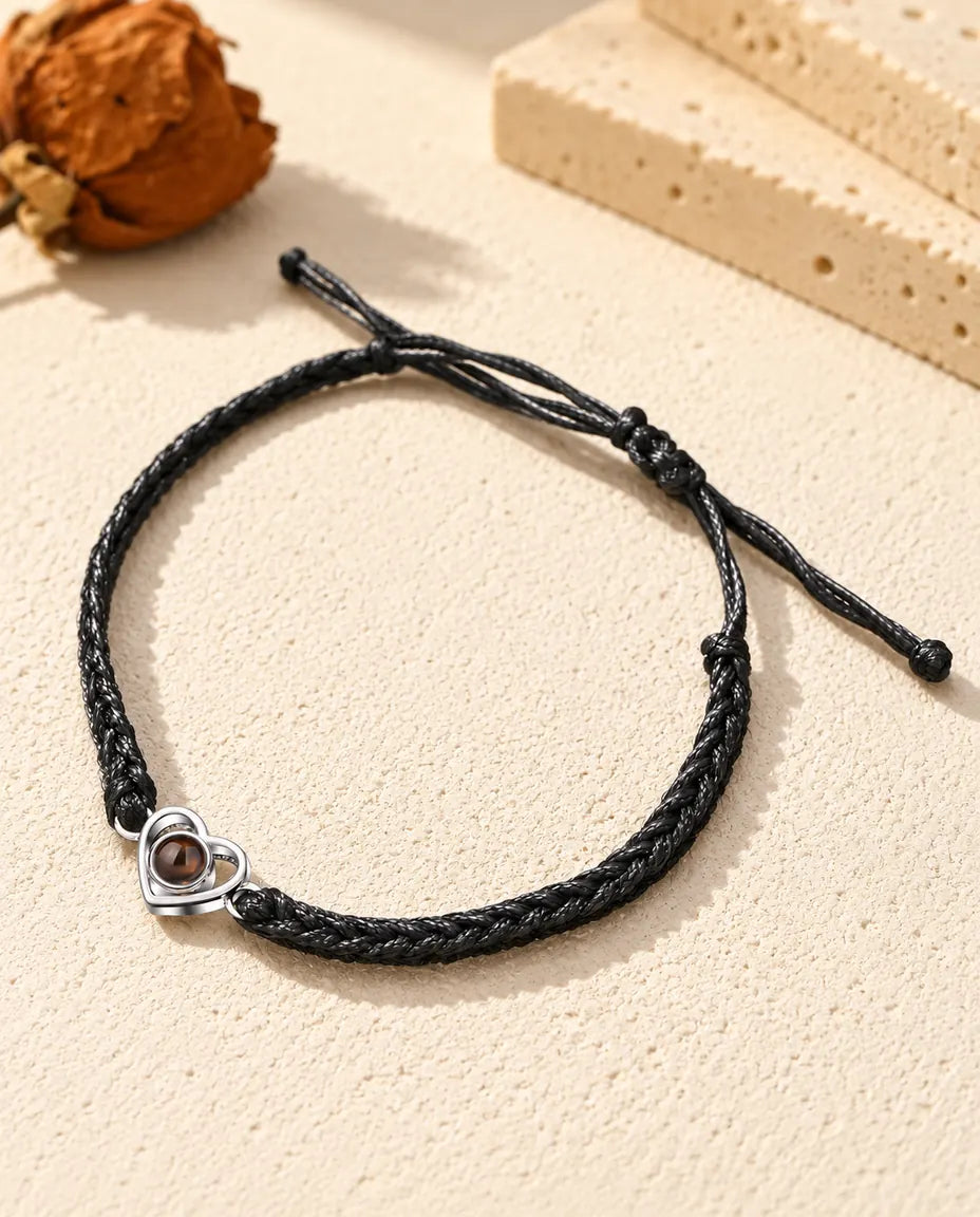 Personalized 925 sterling silver open heart photo projection bracelet on a black braided cord, lying on cream plaster