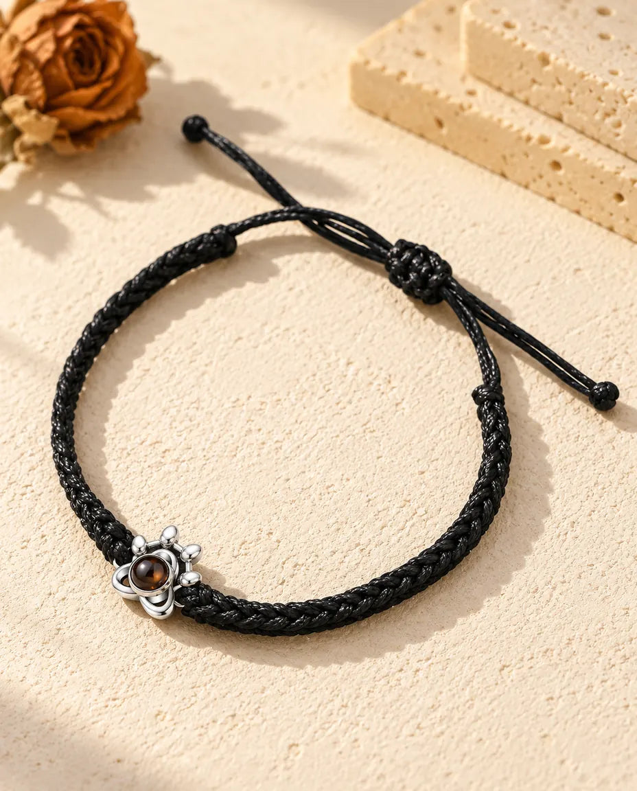 Personalized 925 sterling silver paw charm photo projection bracelet on a black braided cord, lying on cream plaster
